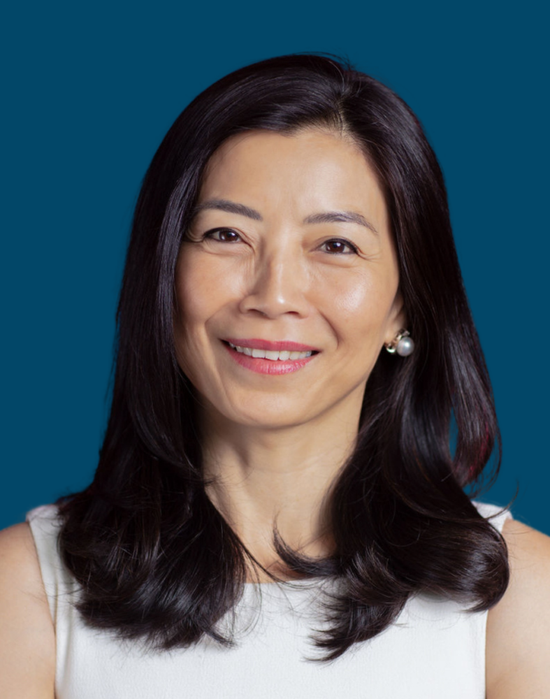 Portrait of Tracy Palandjian, a smiling woman with long, dark hair, wearing a white top, set against a solid blue background.